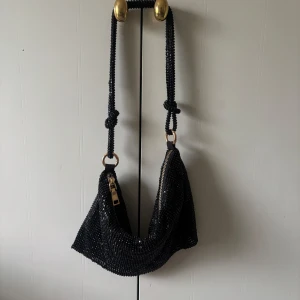 Svart glittrig axelväska - Säljer en svart glittrig axelväska med dragkedja och pärldetaljer. Väskan har en unik form och ett axelband med knutdetaljer. Perfekt för att lyfta din outfit med lite extra bling.
