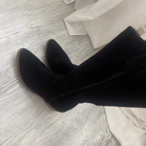 Svarta höga stövlar i mocka - Säljer ett par svarta höga stövlar i mjuk mocka. De har en klassisk form med rund tå och platt sula, perfekta till höst och vinter. Snygga att styla till både kjol och jeans.