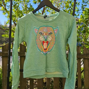 Mintgrön långärmad tröja med tigertryck - Säljer en mintgrön långärmad tröja med ett stort färgglatt tigertryck på bröstet. Tröjan har rund halsringning och är i ett mjukt, tunt material. Perfekt för dig som vill sticka ut med en cool detalj!