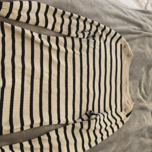 Randig ribbad tröja från H&M med knappar - Säljer en ribbad långärmad tröja från H&M i beige och svart med horisontella ränder. Tröjan har dekorativa silver knappar vid axeln och är i ett stretchigt material. Perfekt för dig som gillar klassisk stil med en twist.