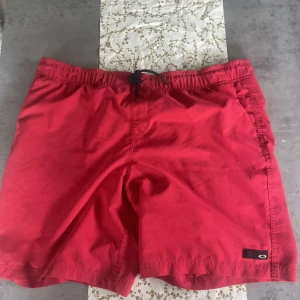 Röda badshorts från Oakley - Snygga röda badshorts från Oakley med elastisk midja och snörning. Klassisk modell med sidofickor och diskret logga på ena benet. Perfekta för sommardagar vid stranden eller poolen.