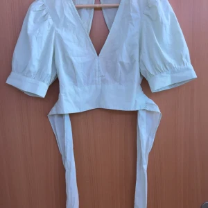 Ljusgrön omlottopp med puffärm från Pieces - Supersöt ljusgrön omlottopp från Pieces med puffiga korta ärmar och djup v-ringning. Toppen knyts i ryggen med långa band och har en snygg rosett bak. Perfekt för att lyfta vilken outfit som helst!