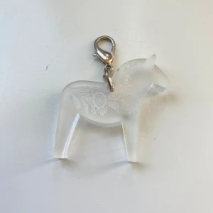 Genomskinlig dalahäst-berlock i akryl - Säljer en söt berlock i form av en dalahäst i genomskinlig akryl med graverat kurbits. Utrustad med en silvrig karbinhake för enkel fastsättning på armband, halsband eller väska.