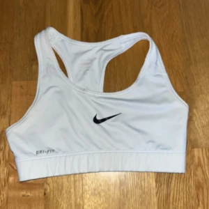 Vit sport-bh från Nike - Säljer en vit sport-bh från Nike med Dri-FIT-teknologi. Modellen har brottarrygg och elastisk nederkant för extra stöd. Perfekt för träning och sportiga aktiviteter. Klassisk Nike-logga framtill.