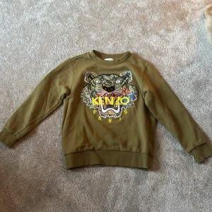 Olivgrön tröja från Kenzo Kids - Säljer en olivgrön tröja från Kenzo Kids med ett stort tigertryck och texten 'Kenzo Paris' i gult på framsidan. Tröjan har långa ärmar och ribbade muddar vid ärmslut och nederkant. 