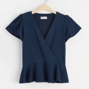 Mörkblå omlottopp från Holly & Whyte - Säljer en mörkblå omlottopp från Holly & Whyte med korta ärmar och volang nertill. Toppen har v-ringning och en feminin siluett som passar perfekt till jeans eller kjol. Det är storlek Xs (Den är stor i modellen och passar xs-m)