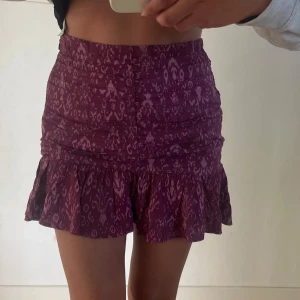 Mönstrad lila kortkjol med volang - Säljer en lila kortkjol med mönster och volang nertill. Kjolen har en figurnära passform upptill och är lätt utsvängd nedtill. 