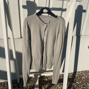 Ljusgrå kofta från H&M - Säljer en ljusgrå kofta från H&M i storlek XS. Koftan har långa ärmar, rund halsringning och knäppning framtill med små knappar. Perfekt att slänga över en topp eller klänning för en enkel och stilren look.