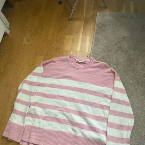 Rosa och vit randig stickad tröja - Mysig stickad tröja i rosa och vitt med breda ränder. Tröjan har rund halsringning och långa ärmar. Perfekt för att hålla sig varm under kyligare dagar.