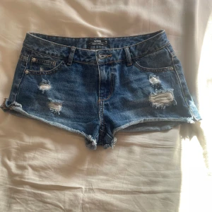 Lågmidjade jeansshorts  - Snygga blå jeansshorts från Sinsay med slitna detaljer och råa kanter. Klassisk femficksmodell med låg midja och knappgylf. Perfekta för varma dagar och en avslappnad stil.