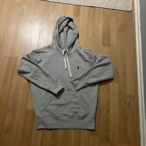 Grå hoodie från Ralph Lauren - Säljer en klassisk grå hoodie från Ralph Lauren med vit dragsko och broderad logga på bröstet. Tröjan har känguruficka och mjukt material, perfekt för en avslappnad stil.