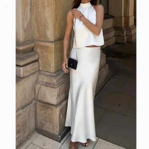 Vit långkjol i satin - Snygg vit långkjol i glansigt satinmaterial med elegant, figurnära passform och matchande topp till. Kjolen har en stilren och minimalistisk design som ger en lyxig känsla. Perfekt för dig som gillar en clean och chic stil. Aldrig använd och säljer då den inte passade mig