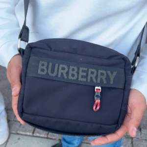 Burberry väska  - Säljer en svart axelväska från Burberry med tydlig logga på både framsidan och axelremmen. Väskan har en dragkedja och justerbar rem, perfekt för att bära det viktigaste. Klassisk och stilren design med Burberry London-etikett inuti.