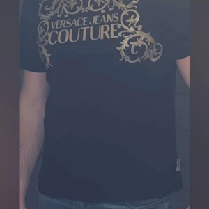 Svart t-shirt från Versace Jeans Couture - Snygg svart t-shirt från Versace Jeans Couture med stort guldfärgat tryck och dekorativa detaljer framtill. Klassisk passform och rund halsringning. 