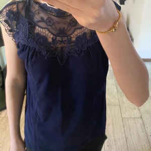 Mörkblå blus med spetsdetaljer - Säljer en elegant mörkblå blus med vacker spets upptill och blommigt mönster. Blusen har ärmlös design och rund halsringning med transparent spets över axlar och bröst.