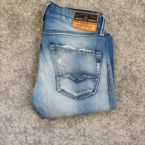 Ljusblå Replay jeans med slitningar - Säljer ett par ljusblå Replay  jeans med snygga slitningar på båda benen och klassiska bakfickor med V-formad söm. Jeansen har en vintage tvätt och läderpatch bak i midjan. Perfekta för en avslappnad stil.storlek 29/32