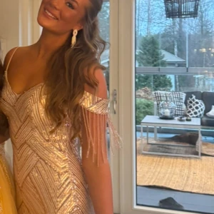Champagnefärgad långklänning med paljetter - Säljer en elegant champagnefärgad långklänning med glittriga paljetter och v-ringning. Klänningen har offshoulder-ärmar med dekorativa fransar och ett geometriskt mönster. Perfekt för bal eller fest.