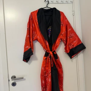 Röd kimono med svarta detaljer och mönster - Säljer en röd kimono med svarta slag och ärmslut. Kimonon har ett vackert asiatiskt mönster med fåglar och blommor samt ett knytband i midjan. Materialet är glansigt och känns lyxigt.