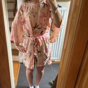 Blommig byxdress med volangärm från D-XEL - Säljer en ljusrosa blommig byxdress från D-XEL med volangärmar och knytskärp i midjan. Det finns dragkedja på baksidan. Stryker innan den postas! Läs gärna bio💘