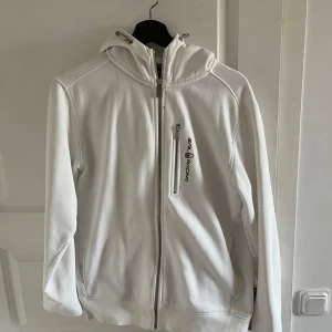 Vit hoodie från Sail Racing - Säljer en stilren vit hoodie från Sail Racing med dragkedja framtill och på bröstfickan. Tröjan har huva och diskret logga på bröstet. Perfekt för en sportig och avslappnad look.