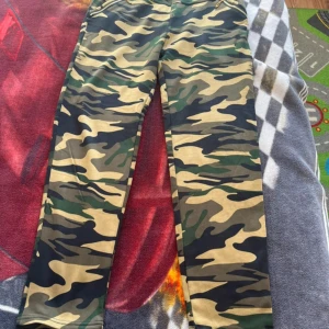 Cargopants i camouflage från RENSANHE - Säljer ett par cargopants i klassiskt camouflage-mönster med gröna, beige och svarta toner. Byxorna har normal passform och är fodrade med mjukt material på insidan för extra komfort. Perfekta för dig som gillar streetwear och vill sticka ut.