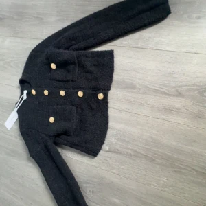 Svart cardigan kofta från Bubbleroom - Säljer en svart cardigan  från Bubbleroom med guldiga knappar och två fickor framtill. Koftan är långärmad och har en mjuk, stickad känsla. Perfekt för lager-på-lager och snygg detalj med de dekorativa knapparna.