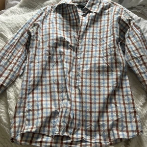 Rutig skjorta från KappAhl i bomull - Säljer en klassisk rutig skjorta från KappAhl i 100% bomull. Skjortan har långa ärmar, knappar framtill och ett mönster i blått, brunt och vitt. Perfekt för dig som gillar en avslappnad men ändå stilren look.