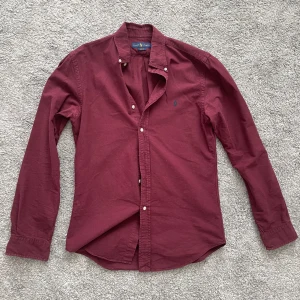Vinröd skjorta från Ralph Lauren - Vinröd skjorta från Ralph Lauren med klassisk button-down krage och broderad logga på bröstet. Skjortan har långa ärmar och vita knappar framtill. Perfekt för en stilren och avslappnad look.