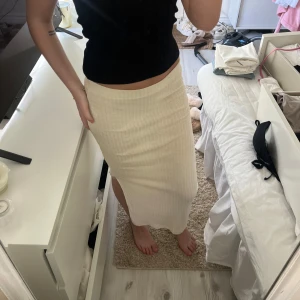 Beige ribbad långkjol - Säljer en beige ribbad långkjol med slits på sidan. Kjolen har en rak passform och är midjehög, perfekt för en stilren och enkel look. Materialet är mjukt och stretchigt för extra komfort.