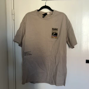 Beige t-shirt från Kodak med tryck - Säljer en beige t-shirt från Kodak med stort färgglatt tryck på ryggen och mindre logga på bröstet. T-shirten har normal passform och är kortärmad. Perfekt för dig som gillar retrostil och grafiska prints.