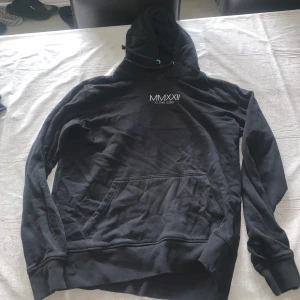 Svart hoodie MMXXII To The Core - Svart hoodie från MMXXII To The Core med vit text på bröstet. Tröjan har huva med snörning och en stor magficka framtill. Perfekt för chill dagar och streetwear-stil. Långärmad och i mjukt bomullsmaterial.