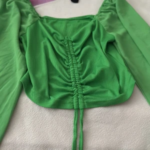 Grön croppad topp med snörning - Snygg grön croppad topp med långa ärmar och cool snörning framtill. Toppen har en ribbad struktur och är tillverkad i ett mjukt syntetmaterial som känns skönt mot huden. Perfekt för dig som vill sticka ut med färg och trendiga detaljer.