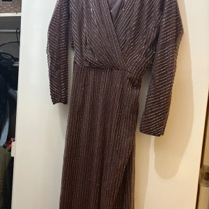 Brun långklänning med pärlor och cutout - Säljer en brun långklänning med glittriga pärlor i rader över hela tyget. Klänningen har lång ärm, omlott upptill och en snygg cutout vid midjan. Materialet är mesh med pärldetaljer som ger en lyxig känsla. Perfekt för dig som vill sticka ut med coola detaljer.
