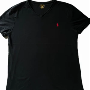 Svart v-ringad t-shirt Polo Ralph Lauren - Svart v-ringad t-shirt från Polo Ralph Lauren med röd broderad logga på bröstet. Slim fit-modell i mjuk bomull som sitter snyggt och känns skön mot huden. Perfekt för dig som gillar stilrena och klassiska plagg med en sportig touch.