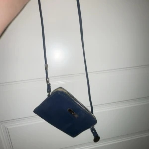 Blå axelväska från Duffy - Snygg liten blå axelväska från Duffy i skinnimitation med justerbar rem. Väskan har ett huvudfack med dragkedja och en mindre ficka med dragkedja på baksidan. Perfekt storlek för mobilen och det viktigaste. Metallplakett med Duffy-logga framtill.