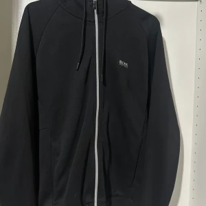 Svart zip-hoodie från Hugo Boss - Svart hoodie från Hugo Boss med dragkedja framtill, snörning i huvan och diskret logga på bröstet och ryggen. Tillverkad i mjuk bomull, perfekt för en chill och stilren look. Snygga detaljer och klassisk passform.