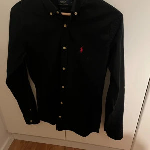 Ralph Lauren skjorta slim fit - Svart långärmad skjorta från Polo Ralph Lauren i slim fit-modell. Klassisk design med knappar framtill och ikonisk röd logga broderad på bröstet. Skjortan är tillverkad i bomull och har en stilren look som passar till många tillfällen. Den är i bra skick och har inga defekter. 