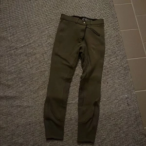 Mörkgröna ridbyxor från CRW, stl 150 - Snygga mörkgröna ridbyxor från CRW i storlek 150. Byxorna har förstärkning på insidan av benen och bak, dragkedjeficka fram och bred resår i midjan. Perfekta för dig som gillar ridning och vill ha en bekväm passform.