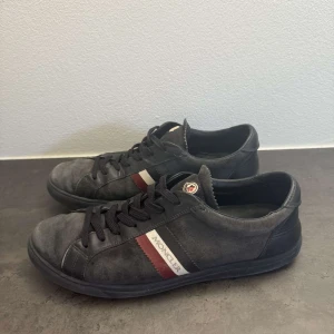 Svarta sneakers från Moncler i mocka - Snygga svarta Moncler sneakers i mocka med svarta snören och klassiska Moncler-loggan på plösen. Sidan pryds av en röd och vit rand med Moncler-tryck. Skorna har rund tå och platt sula, perfekt för en stilren och sportig look.