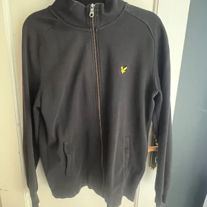 Svart ziptröja från Lyle & Scott - Svart tröja från Lyle & Scott med gul logga på bröstet. Tröjan har hel dragkedja, ståkrage och två fickor framtill. Tillverkad i mjuk bomull, perfekt för en chill och stilren look. Passar om du är 170.