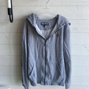 Grå hoodie från Lyle & Scott - Grå hoodie från Lyle & Scott med dragkedja framtill och klassisk gul logga på bröstet. Tröjan har huva, två fickor och är tillverkad i mjuk bomull. Perfekt för alla dagar och årstider och enkel att matcha med jeans eller joggers.