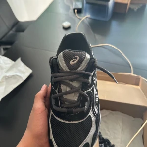 Svarta och grå Asics gel nyc - Säljer ett par snygga Asics sneakers i svart och grått med coola detaljer och meshpaneler för extra ventilation. Skorna har en robust platt sula och klassisk snörning. Perfekt för dig som gillar sportig stil och vill ha bekväma skor till vardagen.