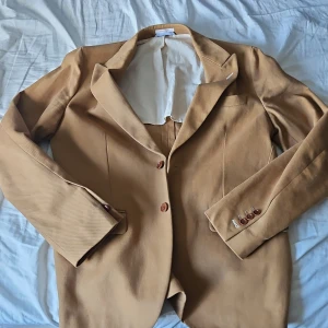 Beige kavaj från GANT Rugger - Snygg beige kavaj från GANT Rugger med klassisk passform och två knappar framtill. Nypris 3000 kr. Kavajen har slät yta, ljus insida och dekorativa knappar vid ärmslut. Perfekt för dig som gillar stilrena och tidlösa plagg med amerikansk sportswear-vibe.