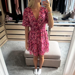 Rosa kortklänning med volanger - Supersöt kortklänning i rosa och lila toner med ett unikt mönster som påminner om leopard. Klänningen har v-ringning, korta puffärmar och smock i midjan för en snygg passform. Volanger i flera lager ger extra flärd och rörelse. Perfekt för dig som gillar färg och mönster!