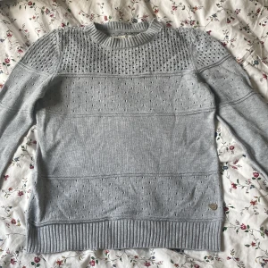Grå stickad tröja från 365 - Snygg grå stickad tröja från 365 med coolt hålmönster upptill och på ärmarna. Rund hals och ribbade muddar vid hals, ärmslut och nederkant. Perfekt för dig som gillar enkel men ändå unik stil. Passar dig som vill ha en chill och bekväm look.