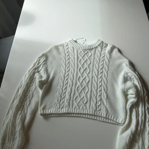 Vit croppad stickad tröja H&M Divided - Säljer en vit croppad stickad tröja från H&M Divided i storlek S. Tröjan har ett snyggt flätstickat mönster framtill och på ärmarna, samt ribbad hals och muddar. Perfekt för dig som gillar trendiga och mysiga plagg med lite edge.
