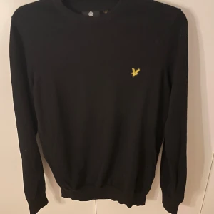Svart stickad tröja från Lyle & Scott - Svart stickad tröja från Lyle & Scott med klassisk rund hals och gul broderad logga på bröstet. Tröjan har långa ärmar och ribbade muddar vid ärmslut och nederkant. Perfekt för dig som gillar stilrena och enkla plagg med snygga detaljer.
