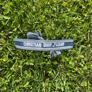 Blått Christian Dior J'Adior armband - Snyggt blått armband från Christian Dior med texten 'J'Adior' broderad i vitt. Armbandet har en justerbar knytning med två tofsar och är gjort i ett mjukt tygmaterial. Perfekt för dig som vill ha en cool designeraccessoar med statement.
