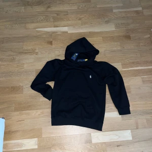 Svart hoodie från Polo Ralph Lauren - Tjena! Jag tänker sälja min hoddie som jag fick i present eftersom jag har fyllt 16. Jag har använt den några gånger och den är Tyvär lite liten på armarna. Den är storlek S                                                                Svart hoodie från Polo Ralph Lauren med klassisk logga broderad på bröstet. Tröjan har huva, magficka och ribbade muddar vid ärmslut och nederkant. Tillverkad i mjuk bomull, perfekt för chill dagar. Snygg och enkel design som alltid funkar.