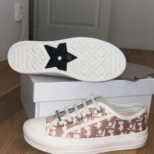 Dior sneakers i rosa och vitt - Snygga Dior sneakers med rosa och vitt monogrammönster, vita snören och platt sula. Skorna har rund tå och tydlig Dior-logga på sidan. Ovandelen är i textil med broderade detaljer och sulan är i vit gummi. Perfekt för dig som vill sticka ut med lyxig streetstyle.